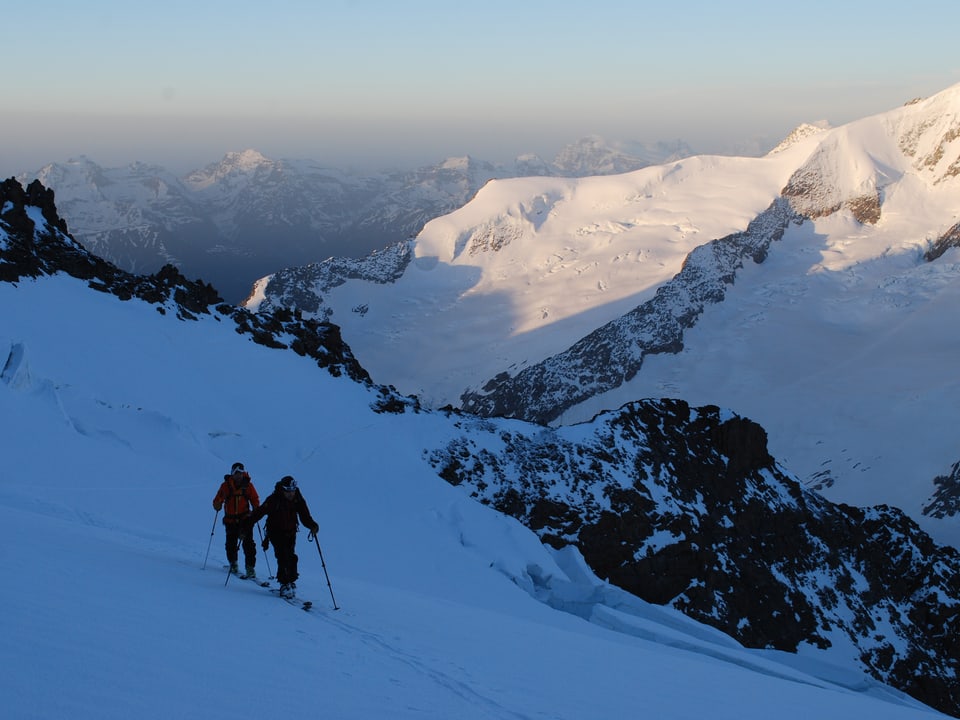 Zwei Männer mit Skiern im Schnee vor Bergkulisse. 