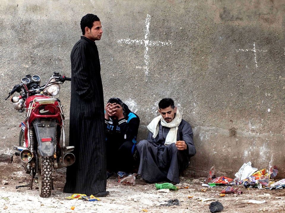 Drei junge Männer sitzen in der ägyptischen Stadt Minya vor einer Hauswand, auf der mit Kreide ein Kreuz gezeichnet wurde.