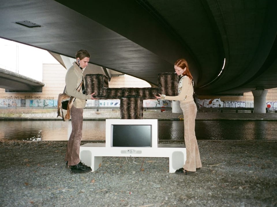 Man sieht einen jungen Mann (links) und eine junge Frau (rechts) mit einer Installation unter einer Brücke.