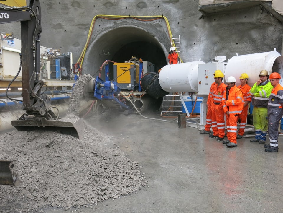 Eine Tunnelbohrmaschine. Daneben stehen ein paar Mineure in orangen Arbeitsanzügen.