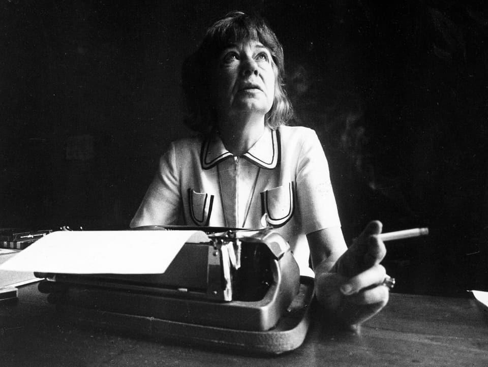 Eine ältere Frau blickt von ihrer Schreibmaschine auf. Sie hält eine Zigarette in der linken Hand.