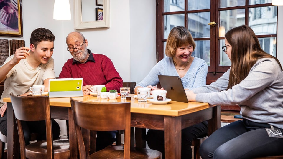 Personen verschiedener Generationen am Tisch. Eine Frau lacht und sieht auf ein Tablet.
