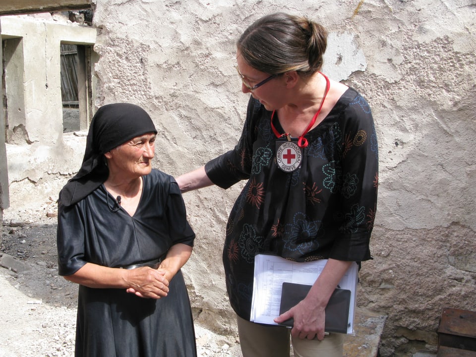 Eine Frau mit dem IKRK-Kreut legt ihre Hand auf die Schulter einer alten, schwarz gekleideten Frau, die besorgt aussieht.