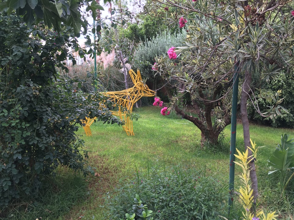 Ein aus Metallstangen geformter, gelber Vierbeiner steht in einem Garten. 