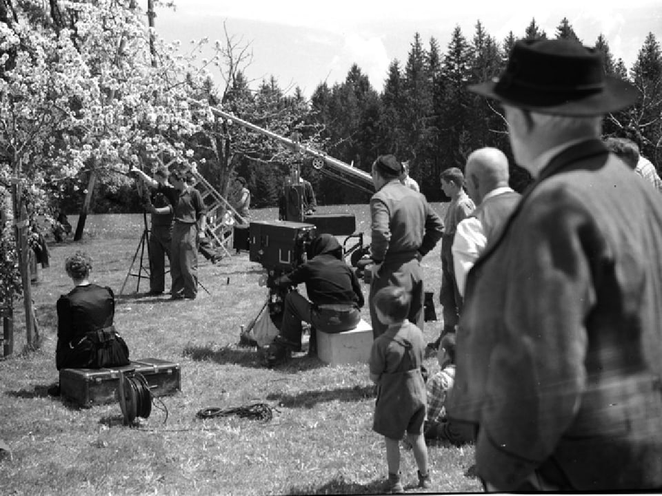 Die Filmcrew auf einer Kirschbaummatte mit blühenden Kirschbäumen. Dazwischen stehen die Kamera, ein Kran und die Schauspieler.