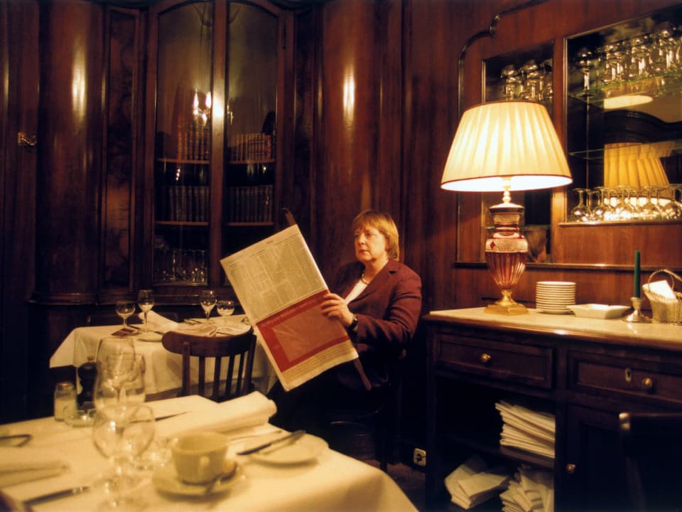 Angela Merkel liest eine Zeitung im schummrig belichteten Café.