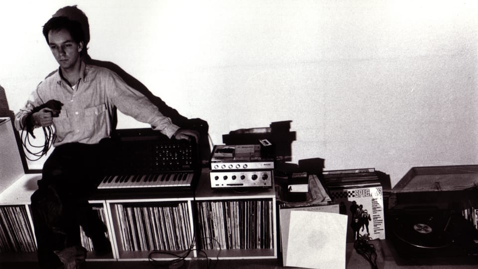 Schwarzweissfoto: Ein junger Mann sitzt auf einem Plattenregal neben einem Synthesizer.