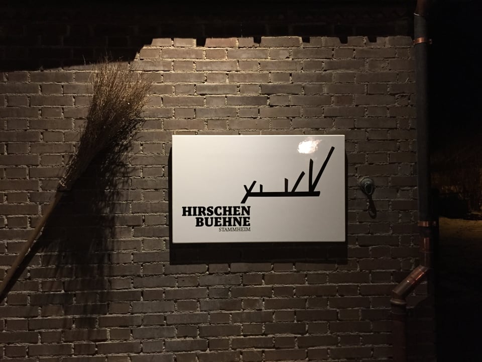 Eine Backsteinwand mit einem weissen Schild, mit schwarzer Schrift das Logo der Hirschenbühne.