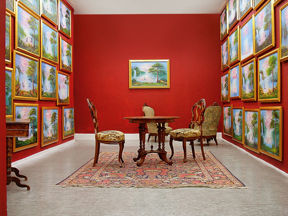 In einem Raum stehen antike Möbel. Die Wände sind leuchtend rot gestrichen. An den Wänden hängen Landschaftsbilder, die sich alle sehr ähnlich sehen. 