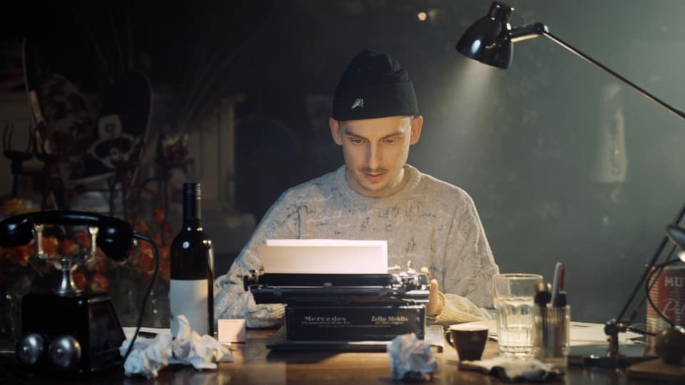 Ein Mann sitzt hinter einer Schreibmaschine am Tisch. Auf dem Tisch steht eine Weinflasche, ein Glas und eine Tasse.