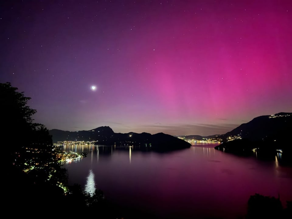 Nächtliche Aussicht auf einen beleuchteten See mit Bergen und rosa Nordlichtern.