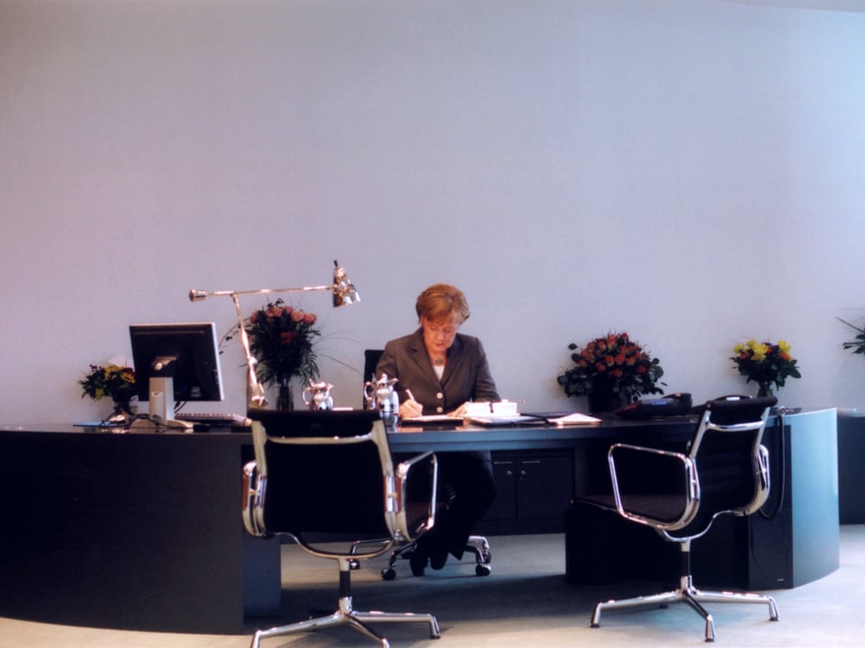 Angela Merkel am Schreibtisch im Büro. Im Hintergund stehen zahlreiche Blumensträusse. 