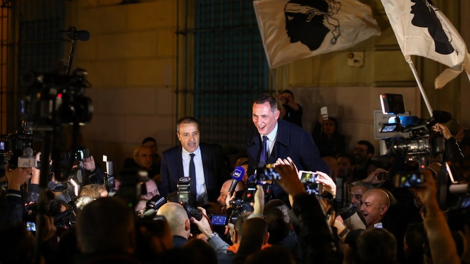 Zwei Männer im Blitzlichtgewitter von Fotografen, es wehen Korsika-Fahnen.