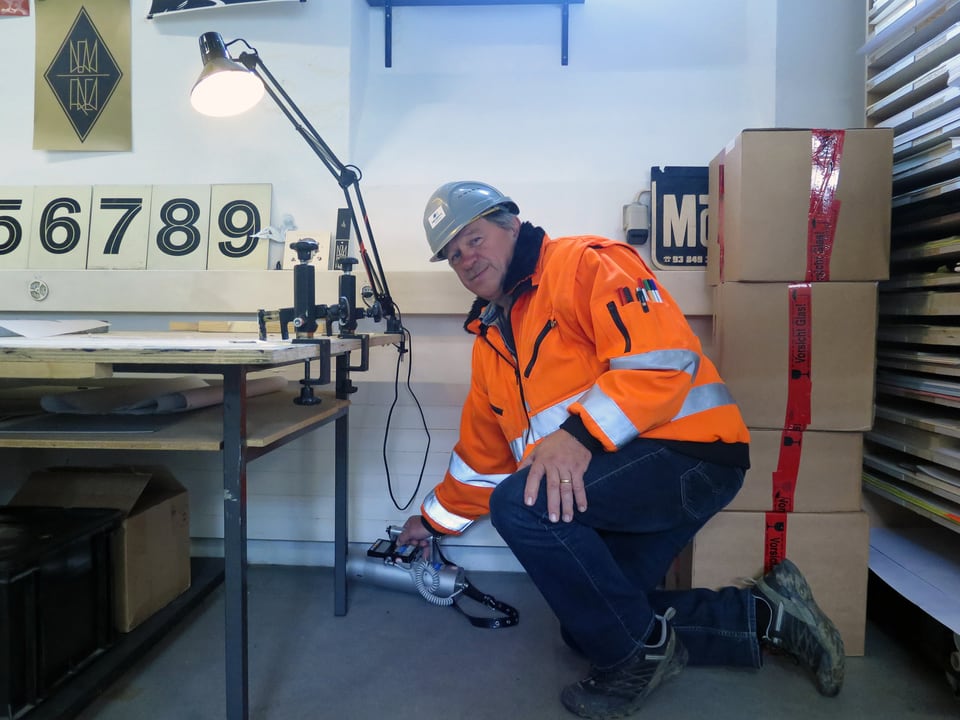 Ein Mann in oranger Leuchtjacke misst mit einem Gerät etwas in einem Arbeitsraum.