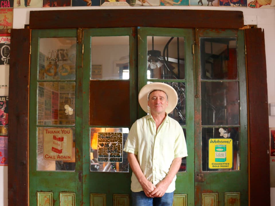 Ein Mann mit Hut steht vor einer grün gestrichenen Türe mit zahlreichen Fenstern. 