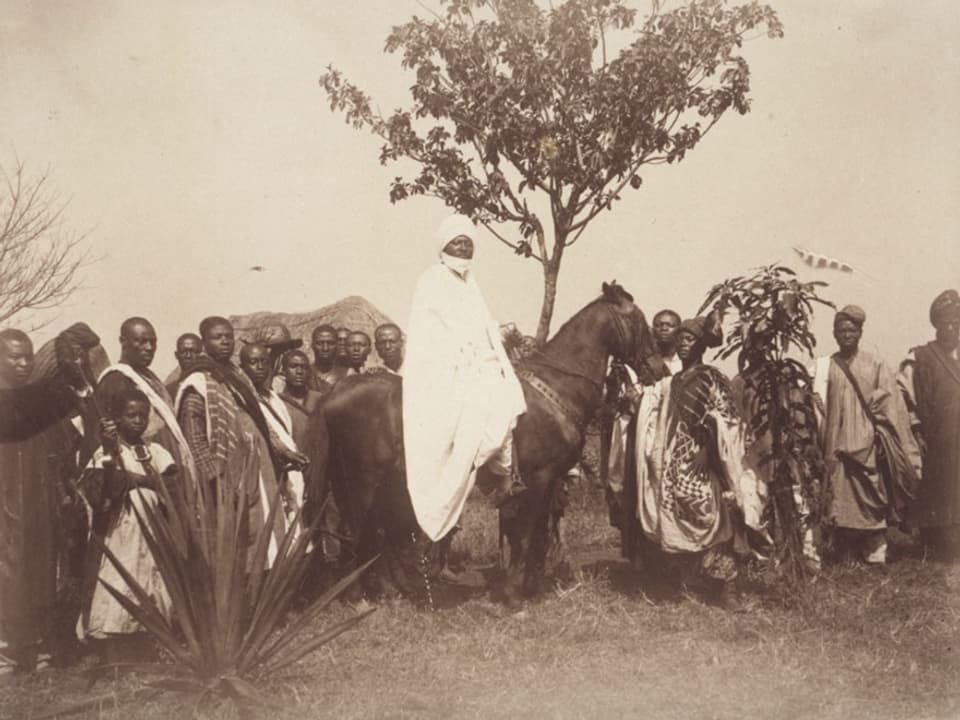 Schwarz-Weiss-Foto: Ein afrikanischer Mann in weissem Gewand sitzt auf einem Pferd, rund um ihn eine Menschengruppe. 