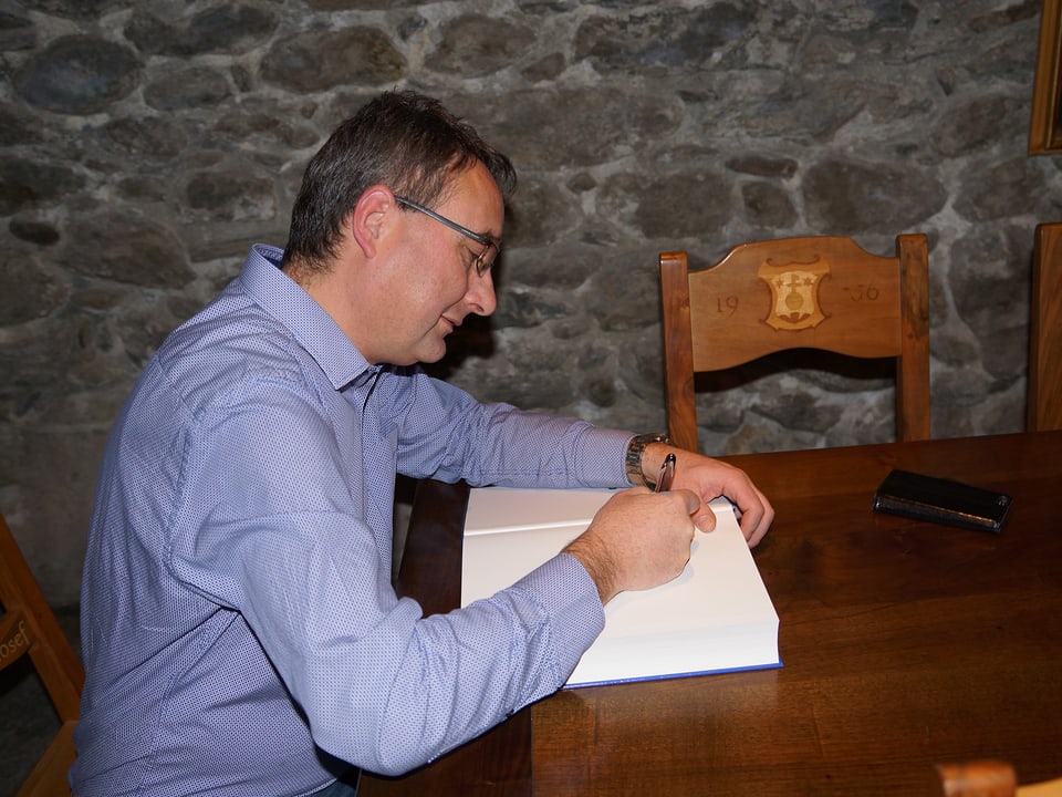 Ein Mann im blauen Hemd sitzt an einem Holztisch und schreibt auf die erste Seite eines dicken Buches.