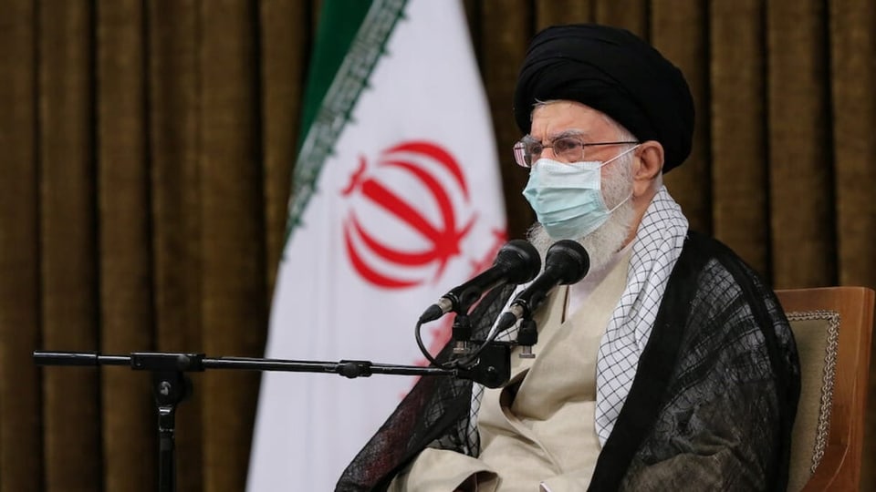 Ayatollah Chomeini trägt eine Gesichtsmaske und spricht in zwei Mikrofone.