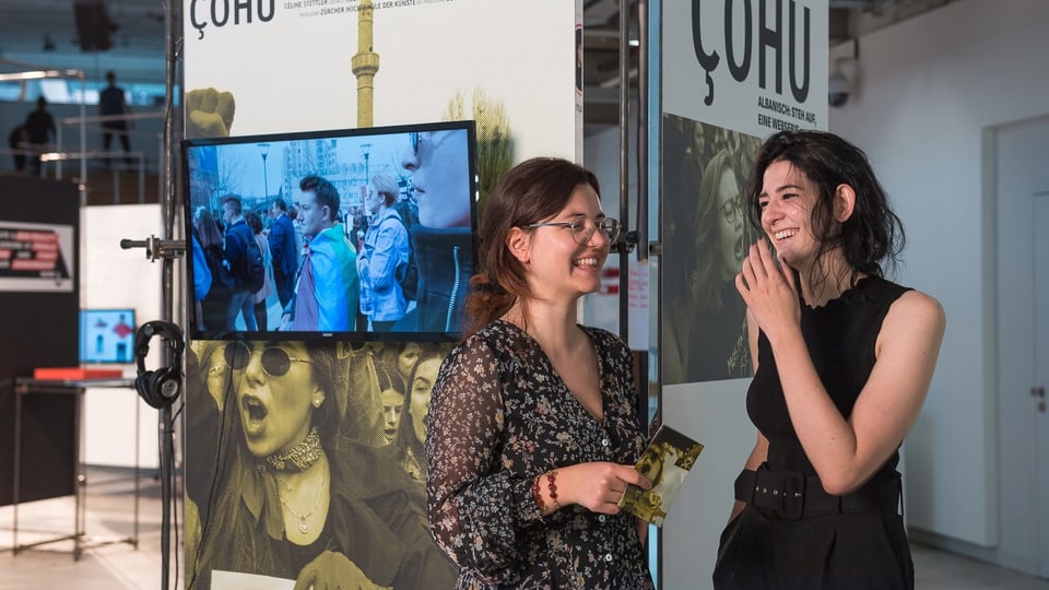 Zwei junge Frauen stehen lachend vor einer Plakatwand mit dre Aufschrift "çohu"