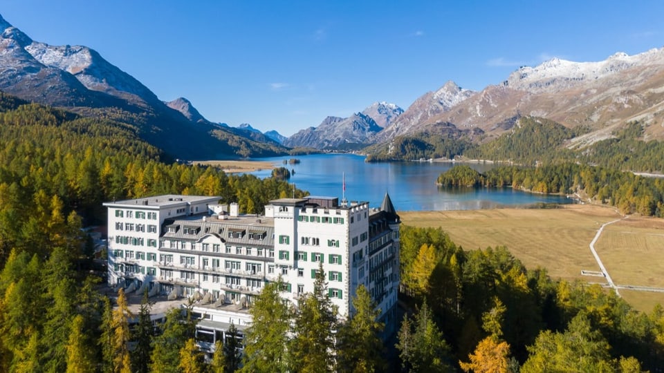 Ein grosses Hotel thront in einem Nadelwald über einem See. Im Hintergrund sind Berge zu sehen.