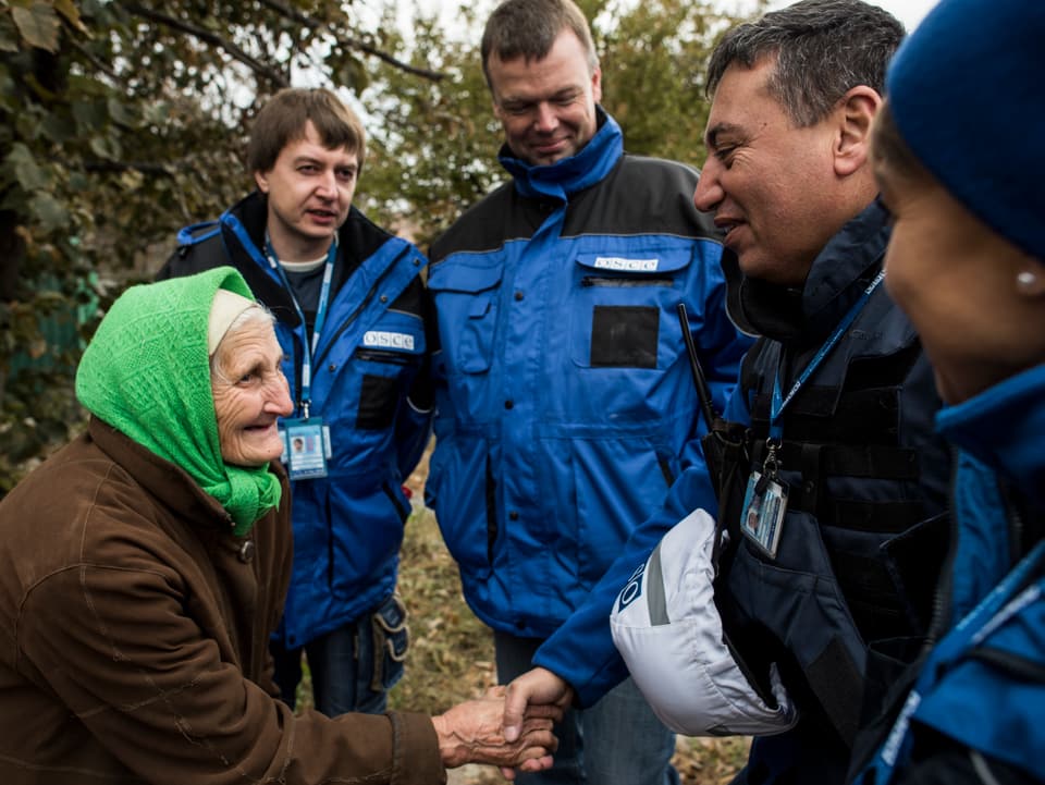 Eine alte Frau mit grünem Kopftuch schüttelt den OSZE-Beobachtern die Hand, sie lächelt.