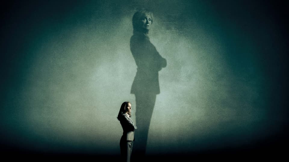 Eine Frau steht in einem dunkeln Raum. Ihr Schatten fällt dramatisch auf die Wand. Auf der Wand ist in dem Schatten wieder ein Frauengesicht zu erkennen.