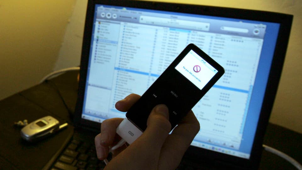 Eine Hand hält einen iPod mit gesperrtem Bildschirm, im Hintergrund ein Laptop mit offenem iTunes-Fenster.
