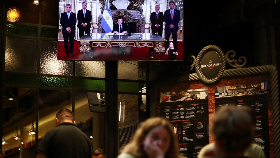Menschen sitzen und stehen in einer Bar. Auf einem Bildschirm ist Javier Milei während seiner Rede zu sehen.