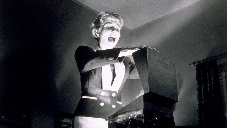 Ein Still aus einem Schwarz-Weiss-Film: Eine Frau öffnet einen Koffer, aus dem Lichtstrahlen kommen.