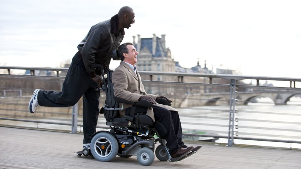 EIn junger, schwarzer Mann steht lachend hinten auf den fahrenden Rollstuhl eines älteren, weissen MAnnes, der ebenfalls lacht.