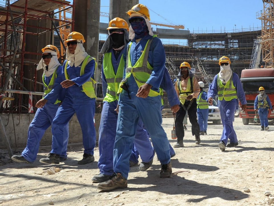 Arbeiter in blauen Arbeitsgewändern und gelben Helmen marschieren auf eine Baustelle.
