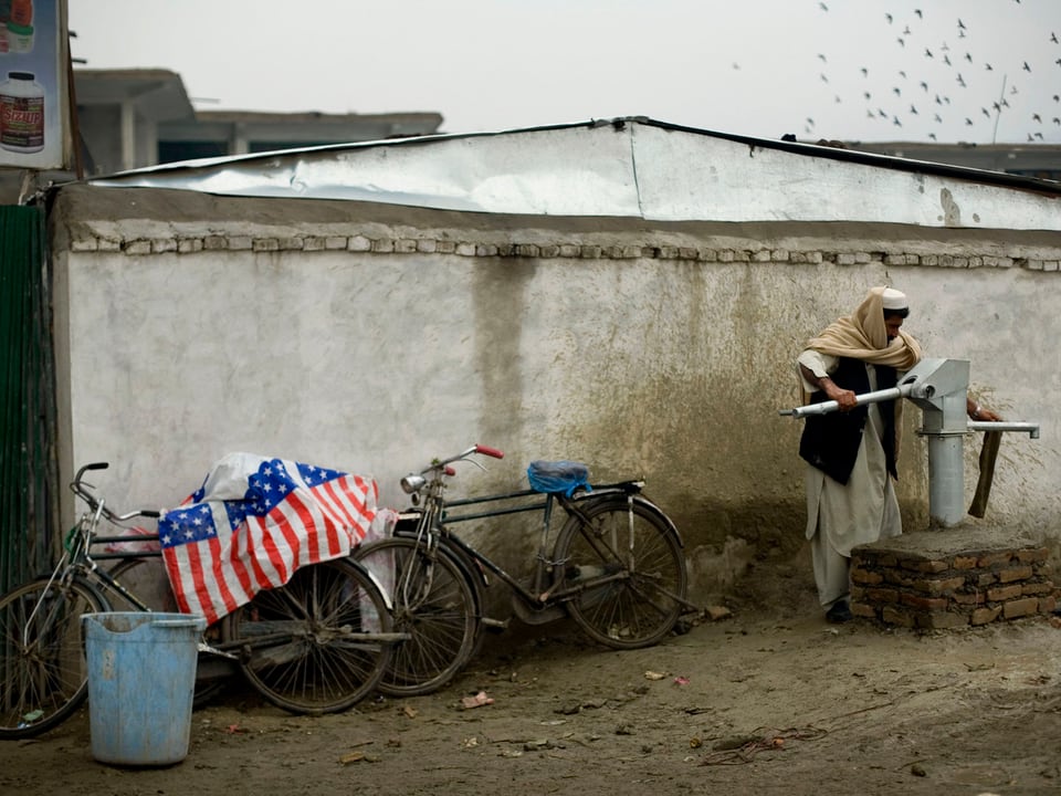 Fahrräder mit Amerika-Flaggen lehnen an einer Wand, daneben bedient ein Mann eine Wasserpumpe