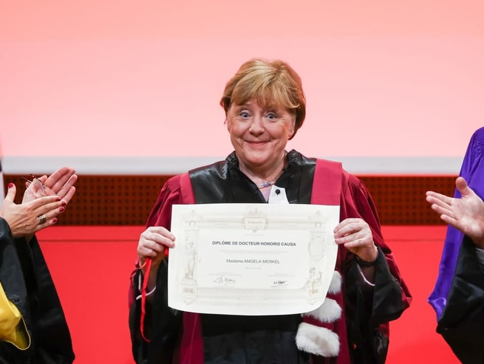 Angela Merkel hält das Diplom in die Kamera und lächelt verkrampft in die Kameras. Sie trägt eine Uni-Kutte.