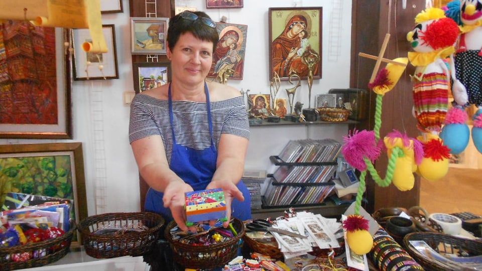 Eine Frau mit blauer Schürze steht in ihrem Laden und hält ein bemaltes Kästchen in der Hand.