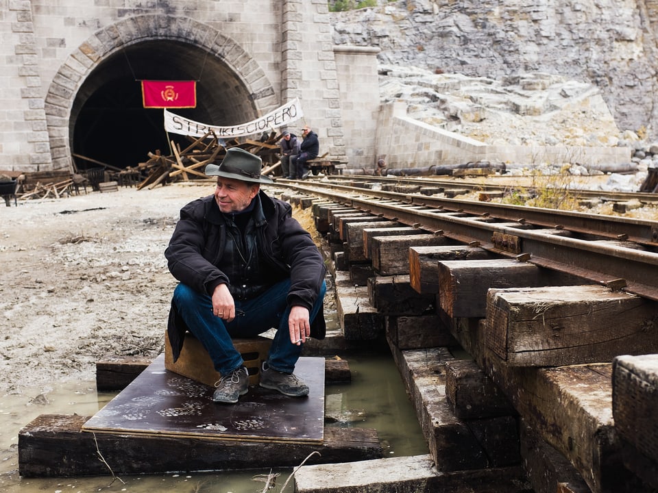 Ein Mann mit Hut sitzt auf einem Holzbrett vor einem Tunneleingang.