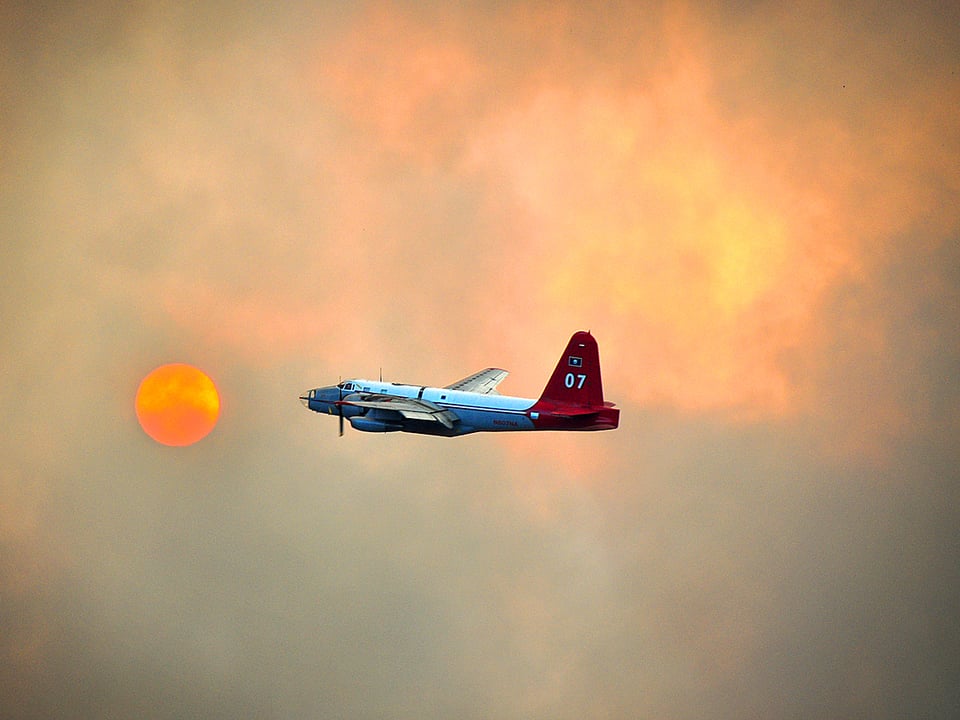 Ein Löschflugzeug kreuzt durch den von Qualm verhangenen Himmel von Texas. Durch den Rauch hindurch ist eine rot glühende Sonne zu sehen.