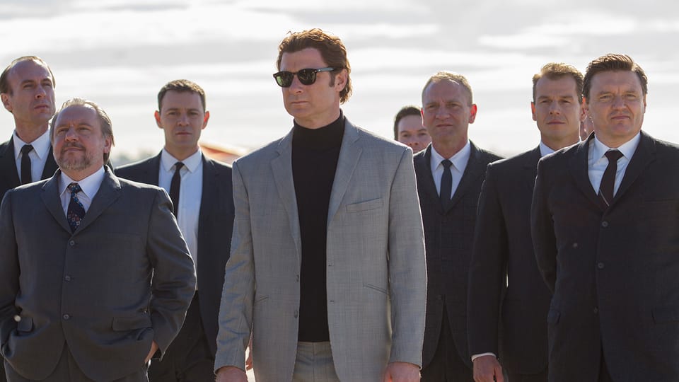 Filmstill: Ein Mann mit Sonnenbrille im hellen Anzug, umringt von dunkel gekleideten Herren.