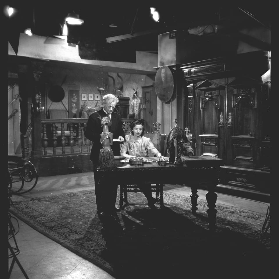 Ein Mann stehend und eine Frau sitzend an einem Tisch. Über ihren Köpfen hängen Filmscheinwerfer.