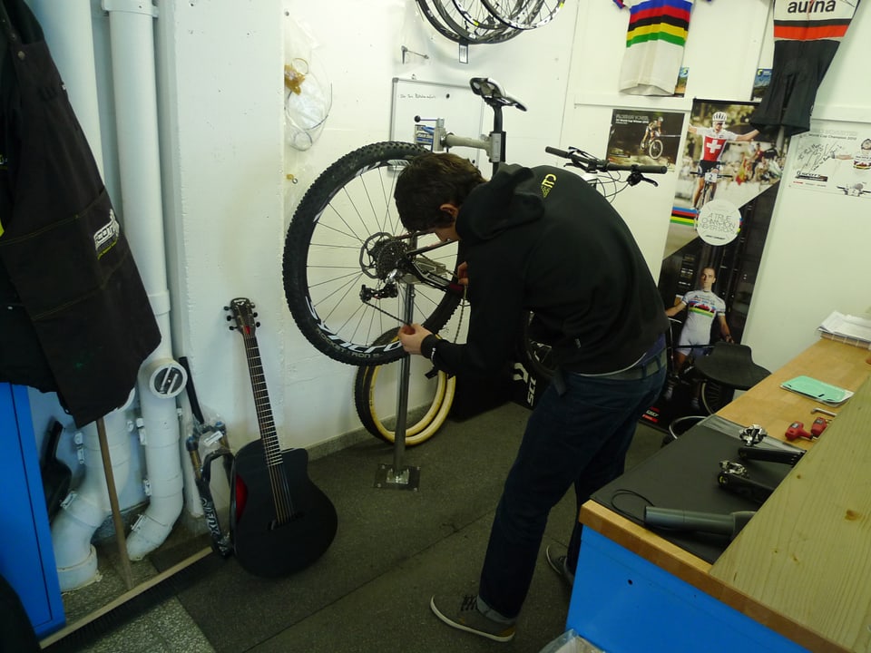 Zu sehen ist ein junger Mann der an einem aufgebockten Moutain-Bike herumrepariert. An der Wand hängen Velo-Trikots und Poster von Velostars. Ausserdem lehnt eine Gitarre an der Wand.