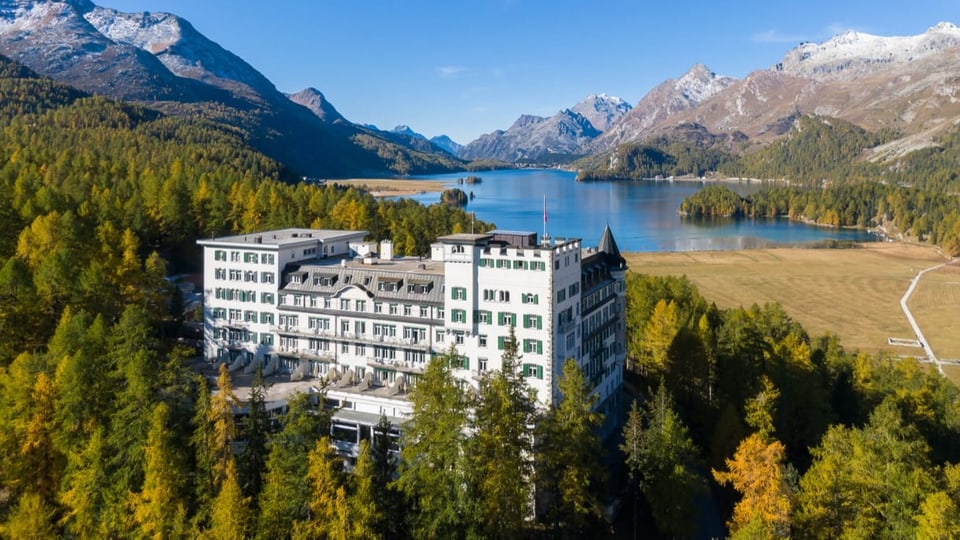 Ein grosses Hotel thront in einem Nadelwald über einem See. Im Hintergrund sind Berge zu sehen.