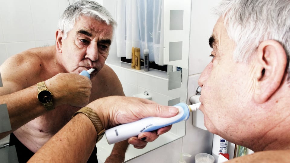 Älterer Mann mit freiem Oberkörper putzt sich im Badezimmer vor dem Spiegel die Zähne und betrachtet dabei sein Spiegelbild.