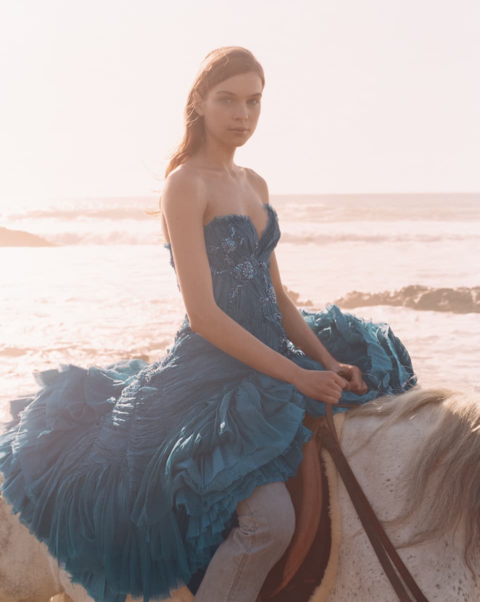 Eine junge Frau sitzt auf einem weissen Pferd, am Strand. Sie trägt ein blaues, wallendes Kleid, da über den Rücken des Pferdes fällt.
