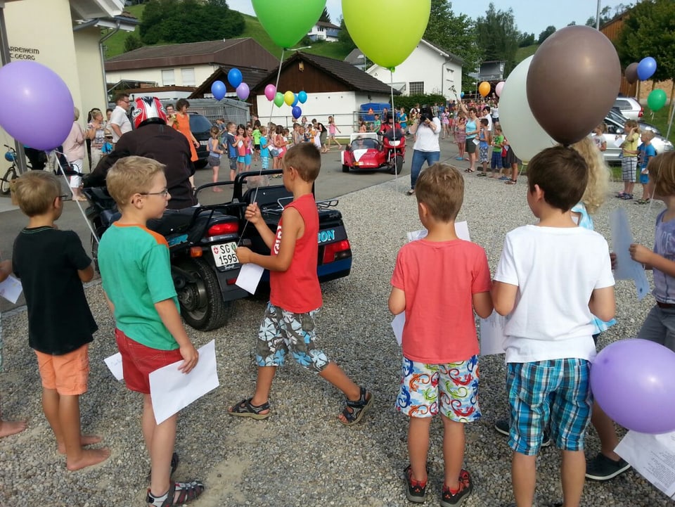 Kinder mit farbigen Luftballons auf einem Dorfplatz.