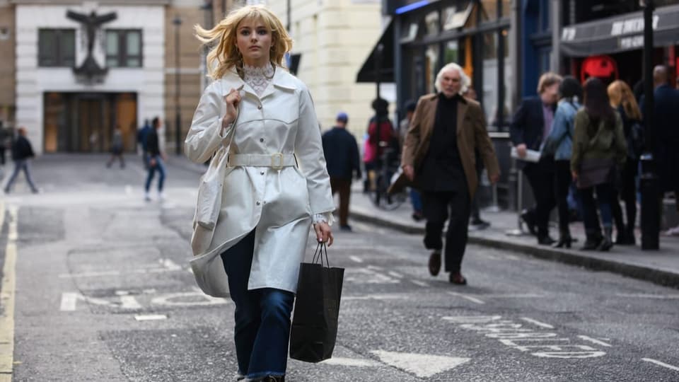 Filmszene: Eine junge Frau mit weissem Mantel geht durch eine Strasse, sie trägt eine Einkaufstüte in der Hand.