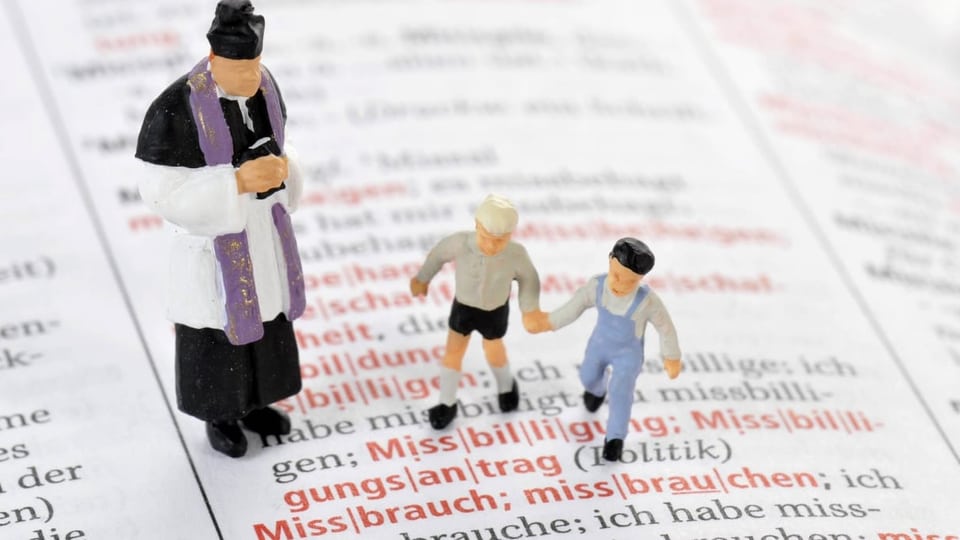 Eine Priesterfigur und zwei Kinderfiguren stehen auf den Worten «Missbrauch» eines Wörterbuchs.