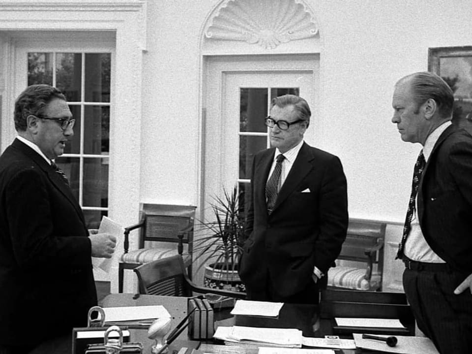 Henry Kissinger steht links und unterhält sich mit Gerald Ford, der rechts hinter einem Tisch steht.