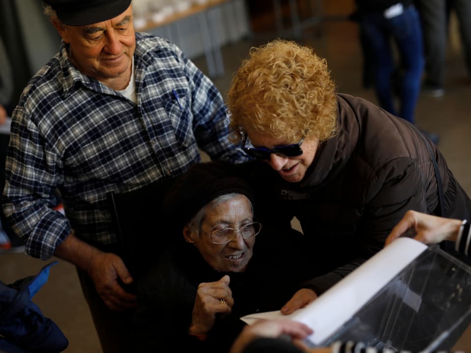 Eine ältere Frau und ein Mann helfen einer Frau im Rollstuhl, ihre Stimme abzugeben.