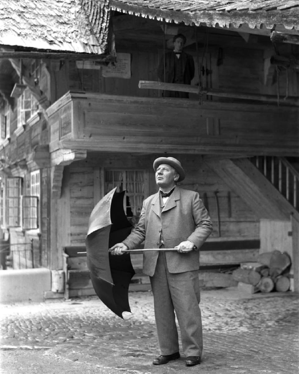 Ein älterer Mann steht vor einem Haus, er öffnet einen Regenschirm und blickt zum Himmel.