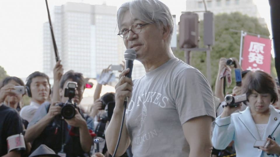 Mann mit runder Brile und grauem Shirt hält Mikro, dahinter Reporter