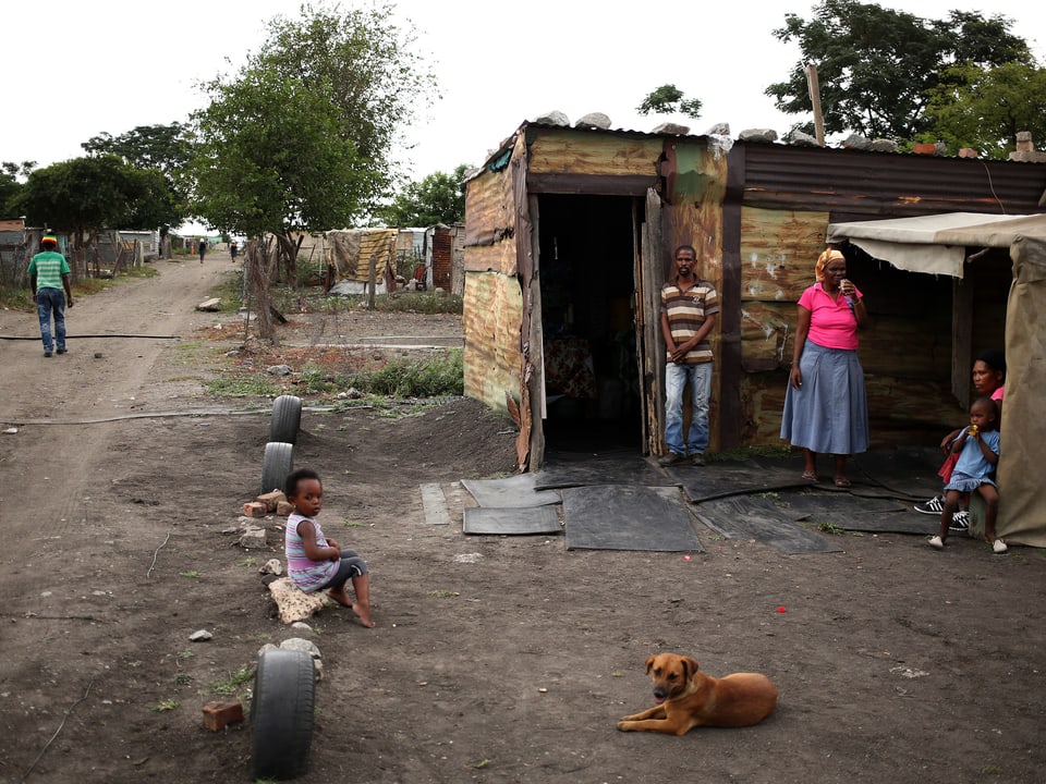 Eltern und drei Kinder sitzen/stehen vor einer Wellblechhütte in einem Township, ein Hund liegt am Boden.